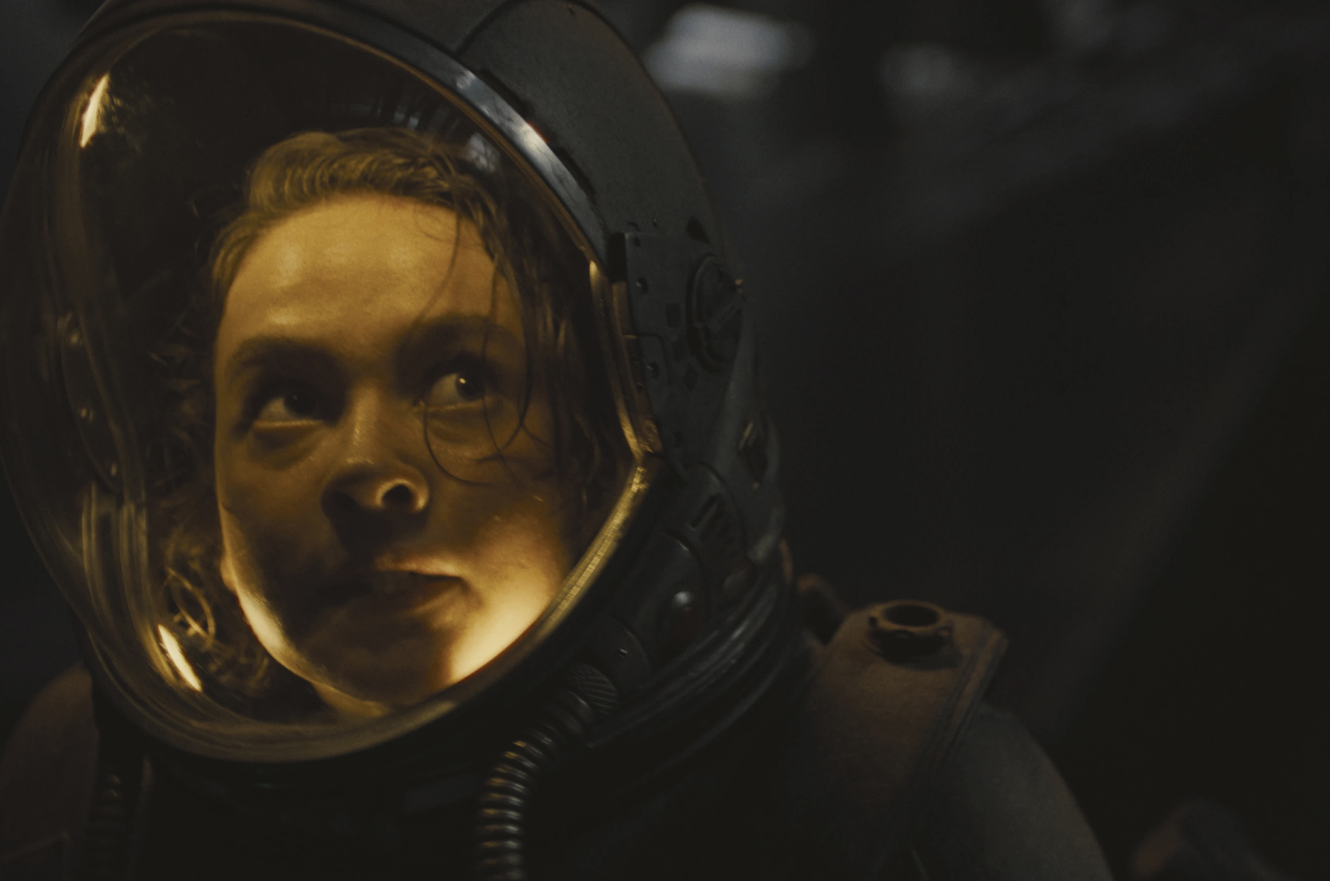 A young woman inside a space suit glances nervously upwards in a gloomy environment.